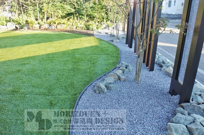 人工芝のエッジ（縁）はお庭の内側から見て柔らかい曲線で描いています。室内からお庭を見ると柔らかい空間となります。樹木の足元には割栗石を置いて、立体感と自然的な空間を演出しています。