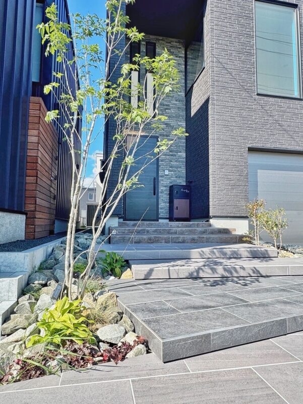 ～植え方でデザインするシンボルツリー～
今回は樹木を少しだけ傾けて浮き階段のステップにかぶさるように植えております。
自然の山であればこのような樹木がたくさん周りにはえており、できるだけ自然風に仕上げたかった狙いがあります！
樹木の足元には割栗石から草花がはえるようなイメージで配置しまして、良い感じに仕上がっています！！
手前に大きい樹木を植え、奥に低木を植えることで奥行感が出せる効果があります。